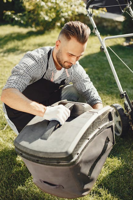 A young man with styled brown hair and a beard, wearing a checkered shirt and black apron, is crouching on a well-maintained lawn during daytime. He is inspecting or adjusting a large, black outdoor g