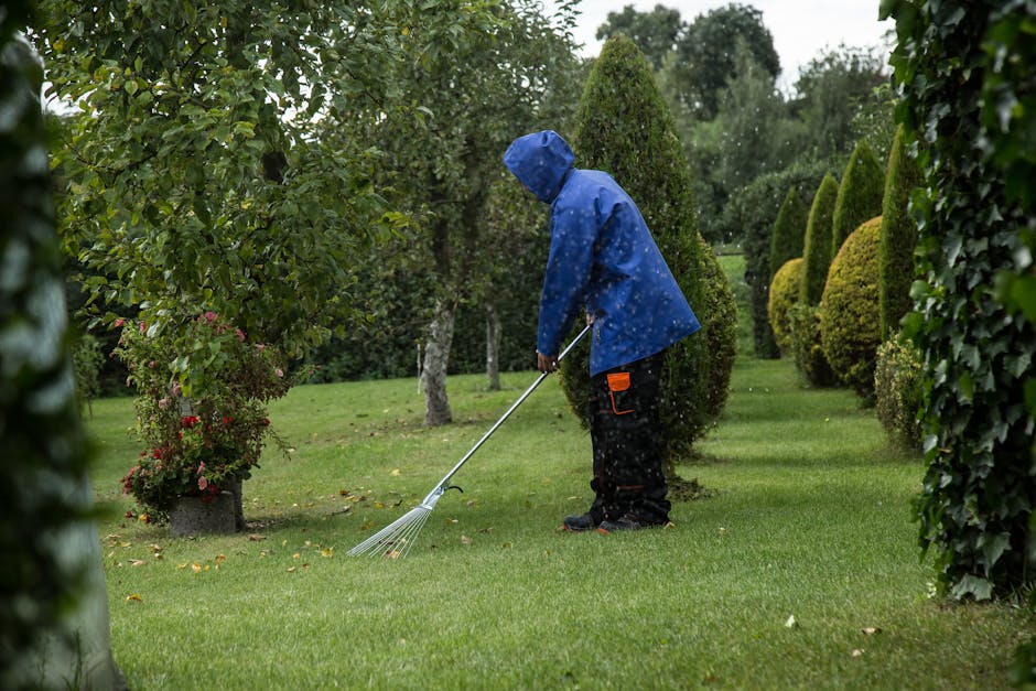 A young man with styled brown hair and a beard, wearing a checkered shirt and black apron, is crouching on a well-maintained lawn during daytime. He is inspecting or adjusting a large, black outdoor g